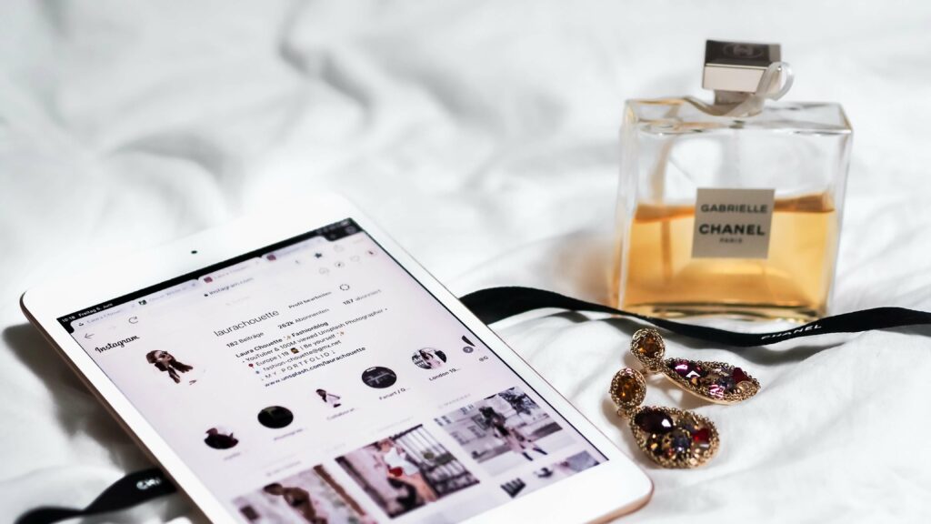Flacon de parfum Gabrielle Chanel, boucles d’oreilles élégantes et profil Instagram sur tablette posés sur un lit blanc